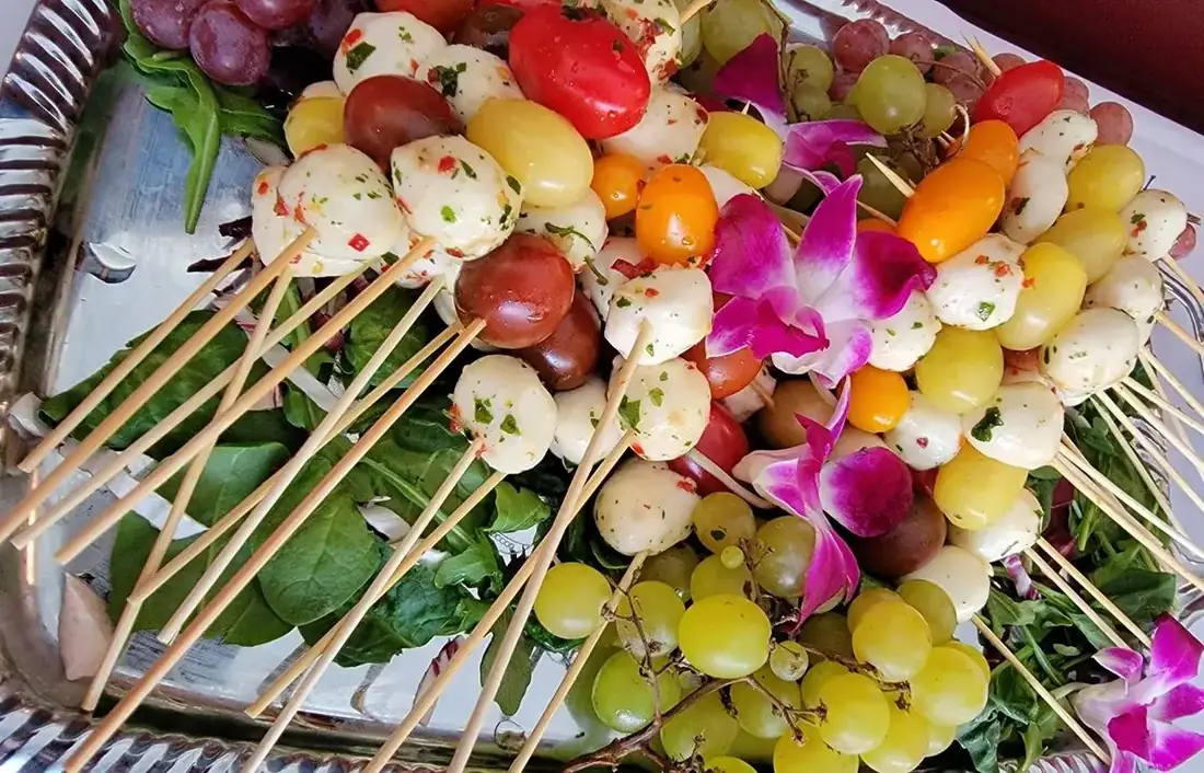 Caprese skewers with marinated mozzarella and heirloom tomatoes arranged on a silver tray with grapes and orchids.