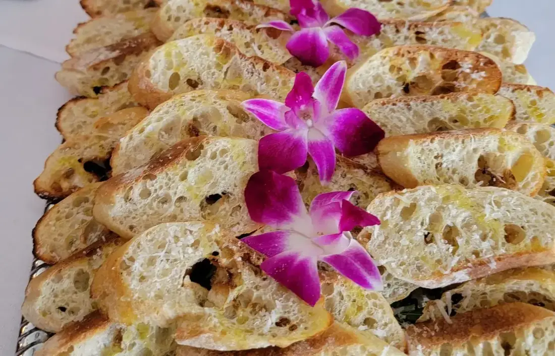 A long silver tray of grilled crostini dressed with purple orchids, beside hummus, crudité and a bowl of bruschetta.