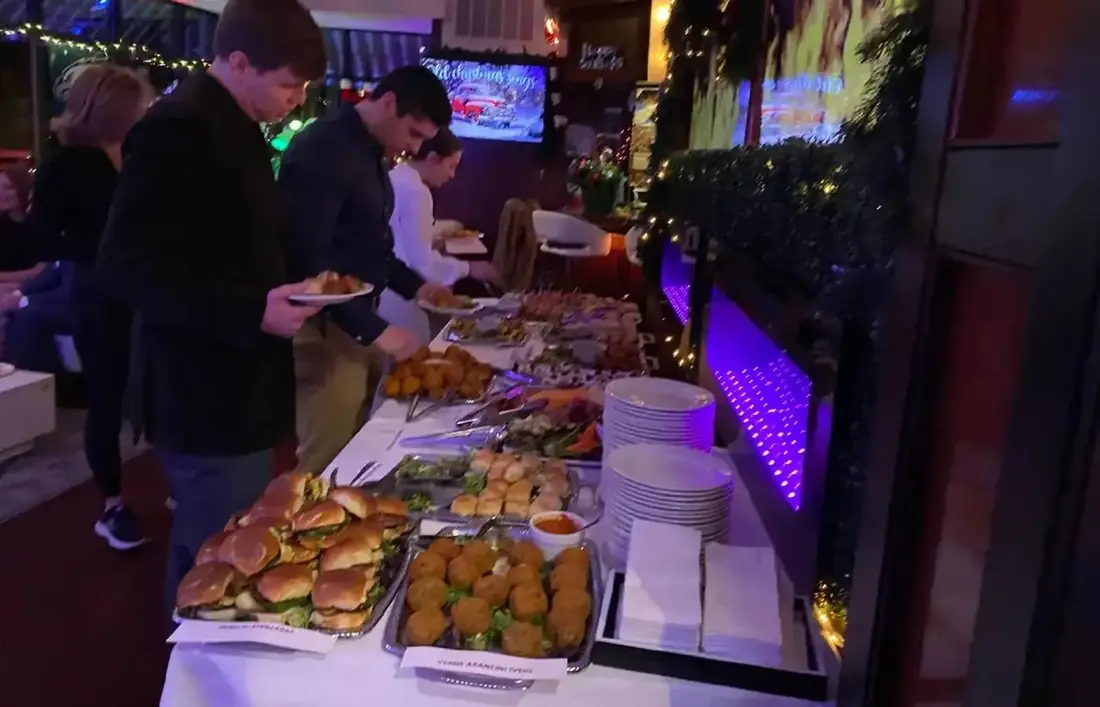 A labelled catering buffet at a corporate holiday party — short rib sliders, verde arancini marked vegetarian, and passed skewers along a white-linen table.