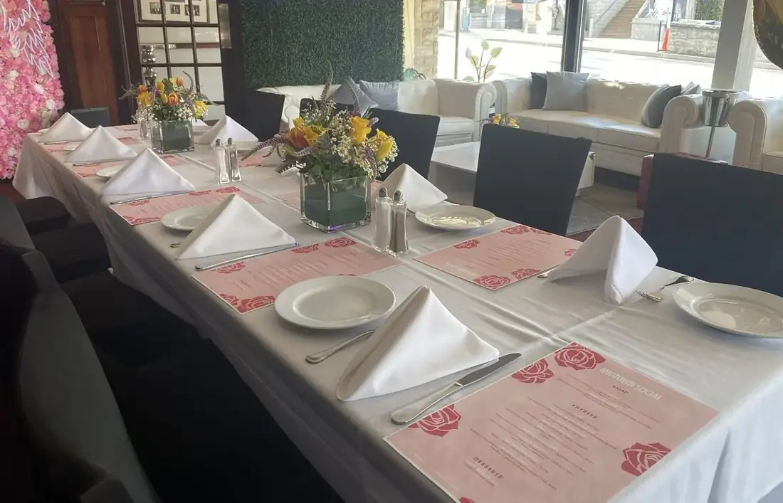 A bridal shower table set with white linen, pink rose menus and flowers under the Midtown Social Lounge and Events neon sign.