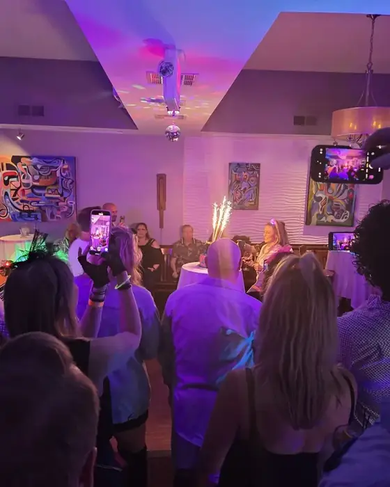 Guests gathered around a birthday cake with sparkler candles, phones raised.