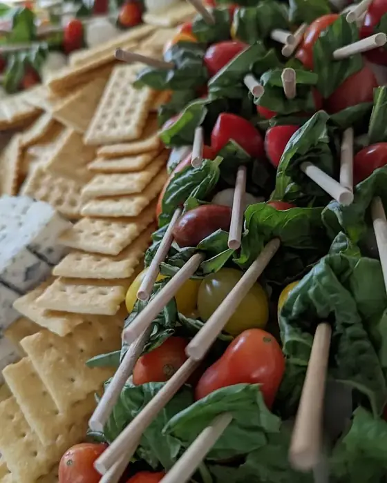 Caprese skewers with basil and heirloom tomatoes beside blue cheese and crackers on a catering board.