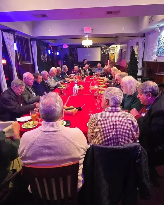 A long table of guests at a club dinner on the first floor with name badges and a projection screen.