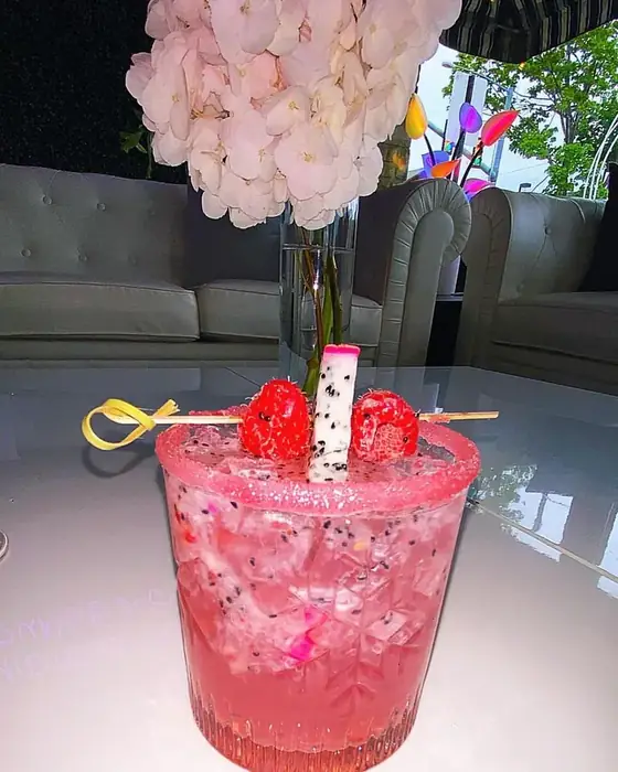 A pink cocktail garnished with dragon fruit and raspberries on a table with white hydrangeas.