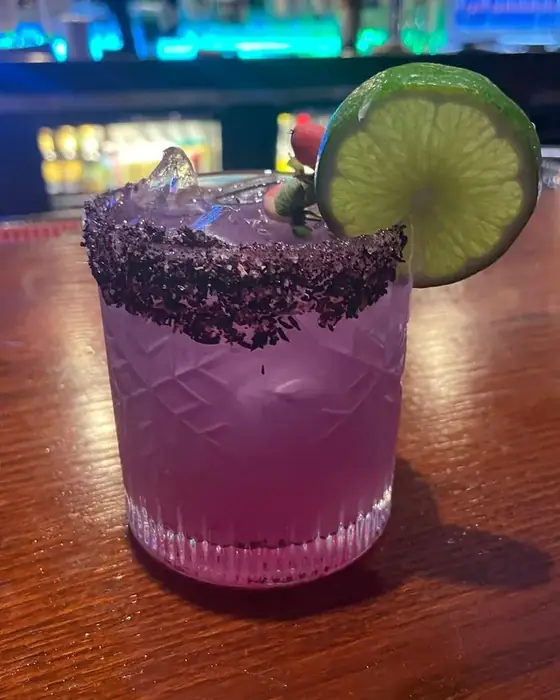 A violet cocktail with a hibiscus salt rim and lime wheel on the bar.