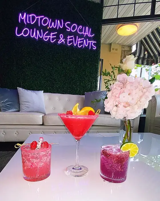 Three signature cocktails on a white table beneath the Midtown Social Lounge and Events neon sign, with hydrangeas.