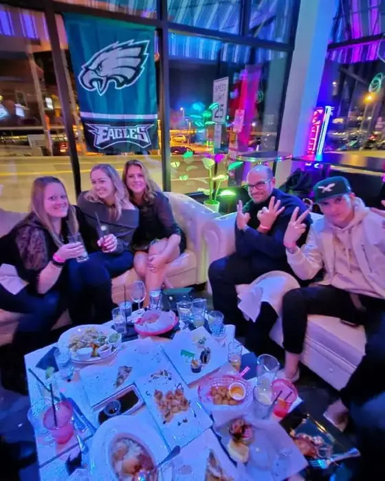 Friends on the white sofas with a spread of food and drinks, Eagles flag in the window behind.