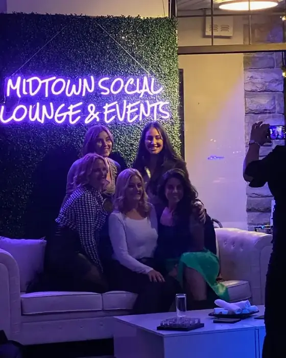 A group of friends on the white lounge sofas in front of the Midtown Social Lounge and Events neon sign.