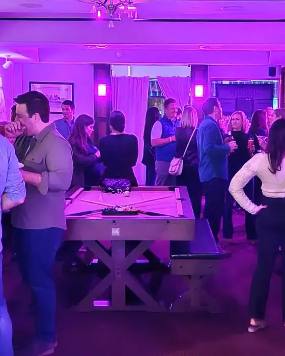 Guests standing around the pool table in the front room under purple lighting.