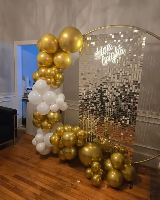 A gold and white balloon arch beside a silver sequin shimmer wall with a shine bright neon sign.