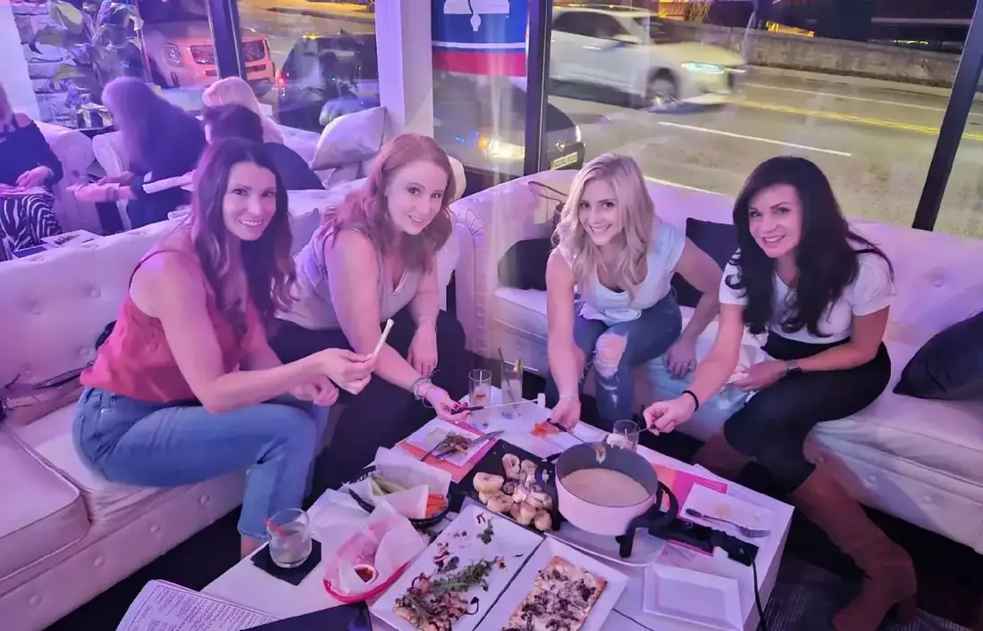 Four friends on a white sofa at Midtown Social sharing fondue and flatbreads, with Lancaster Avenue through the window behind.