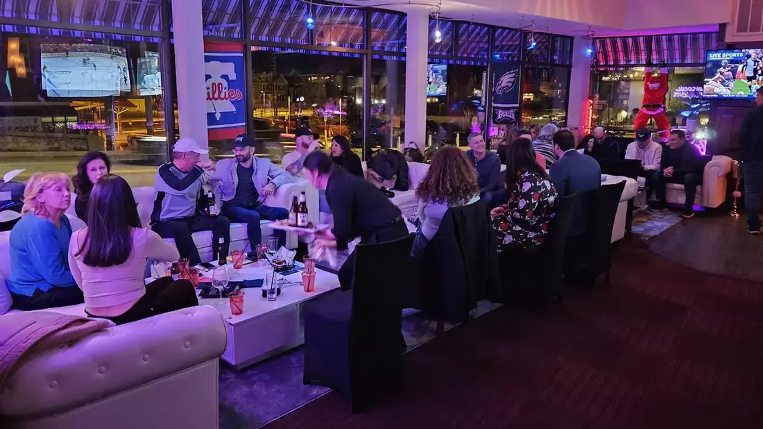 The first floor lounge at Midtown Social on a busy night — white sofas full of guests, hockey and UFC on the big screens, Phillies and Eagles flags in the windows, Lancaster Avenue outside.