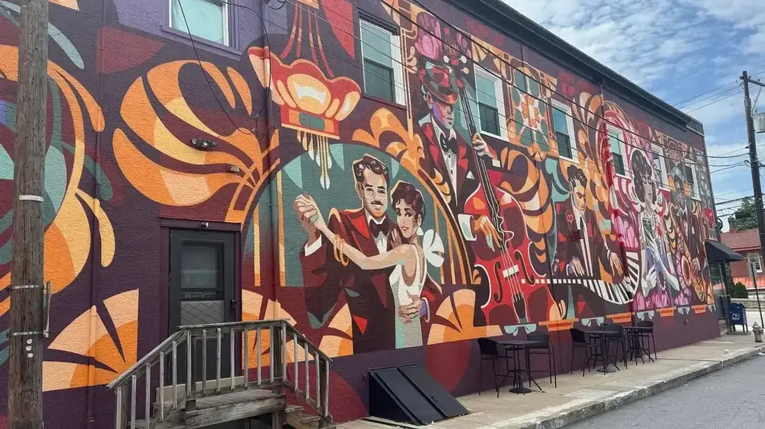 A large mural along the Main Avenue side of the Midtown Social building in Berwyn, painted in purple, orange and teal, showing a couple dancing, a double bass player in a hat, a pianist and singers among art deco shapes.