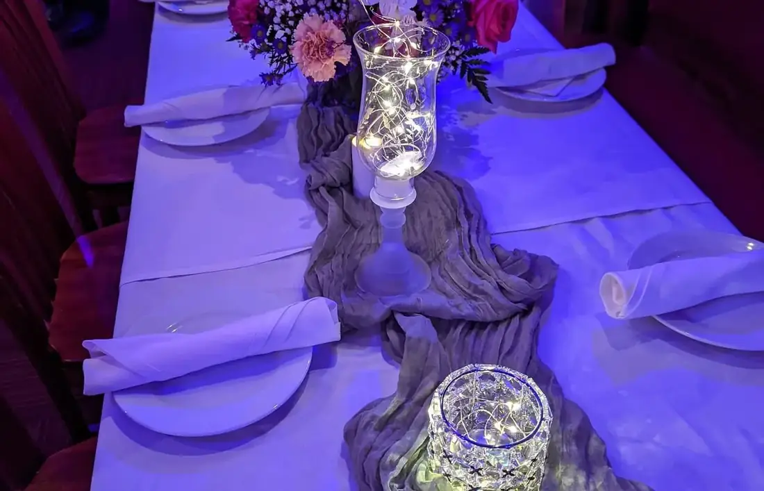 A long table styled with white linen, a soft cheesecloth runner, fairy lights in glass hurricanes and a pink and blue floral centrepiece.